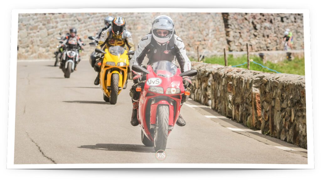 Ein rotes Motorrad wird von einem Fahrer mit Schutzkleidung und Helm gesteuert, während zwei weitere Motorräder im Hintergrund fahren. Die Szene spielt sich auf einer kurvenreichen Straße mit einer Steinmauer am Rand ab, umgeben von einer grünen Landschaft.