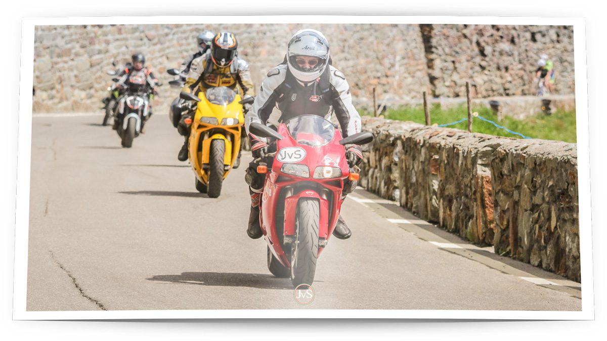 Ein rotes Motorrad wird von einem Fahrer mit Schutzkleidung und Helm gesteuert, während zwei weitere Motorräder im Hintergrund fahren. Die Szene spielt sich auf einer kurvenreichen Straße mit einer Steinmauer am Rand ab, umgeben von einer grünen Landschaft.