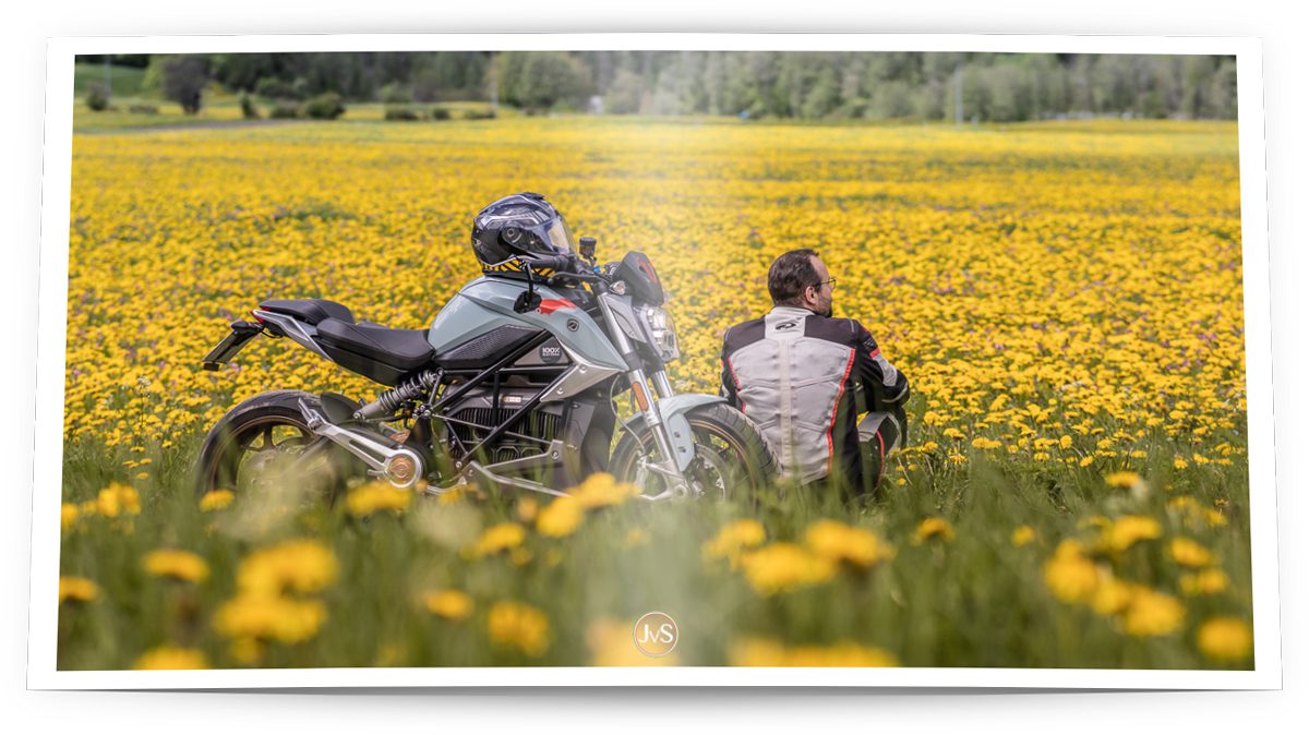 Ein Motorradfahrer sitzt im Grünen auf einer Wiese voller Löwenzahn genießt die Aussicht. Neben ihm steht ein modernes Motorrad.