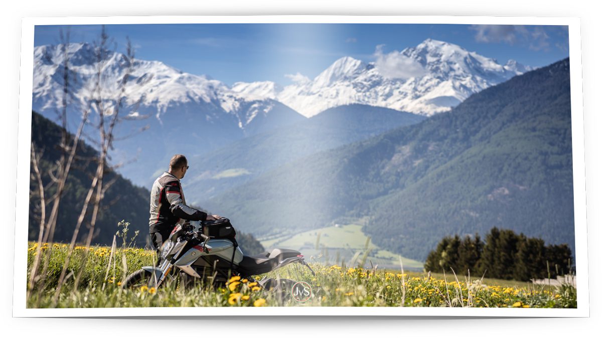Ein Mann, der hinter einem Motorrad in einem Feld mit gelben Blumen steht und auf eine schneebedeckte Berglandschaft schaut.