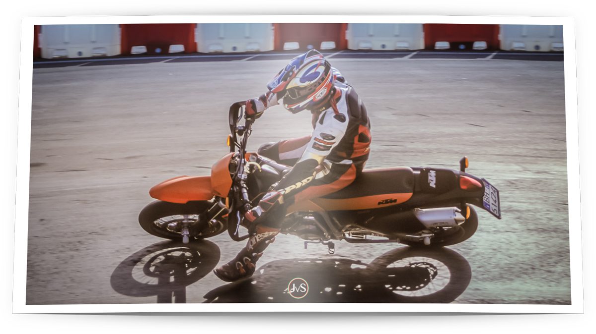 Ein Fahrer in voller Schutzkleidung bewegt eine KTM Supermoto dynamisch durch eine enge Kurve auf Asphalt. Die Szene vermittelt Geschwindigkeit und Fahrfreude, betont durch die Schräglage des Motorrads und die konzentrierte Körperhaltung des Fahrers.