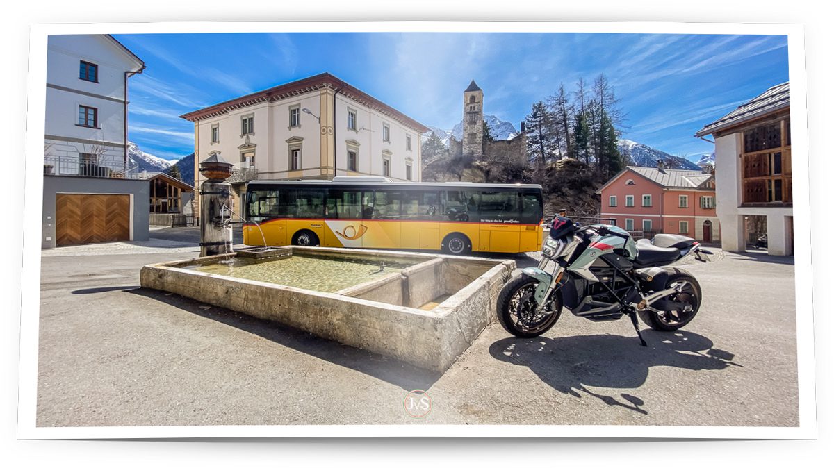 Ein sonniger Platz in einer kleinen Ortschaft im Unterengadin. Im Vordergrund steht ein graues Elektromotorrad neben einem Brunnen, dessen klares Wasser ruhig in das rechteckige Becken fließt. Im Hintergrund parkt ein gelber Schweizer Postbus vor einem eleganten historischen Gebäude. Dahinter ragt der Kirchturm einer alten Dorfkirche in den blauen Himmel, eingerahmt von schneebedeckten Bergen und einigen Bäumen.