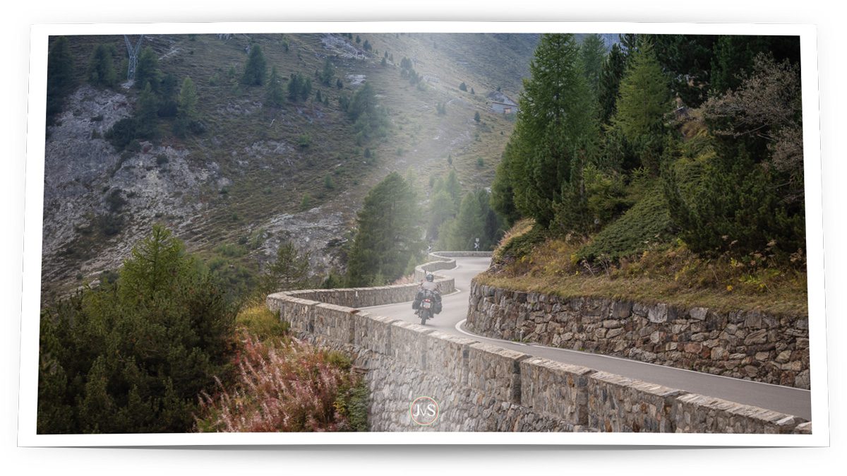 Ein Motorradfahrer, unterwegs auf einer kurvigen Pass-Strasse, begrenzt von Stützmauern aus Naturstein.